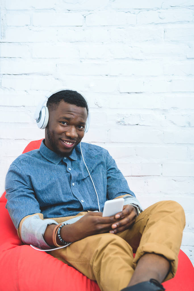 Handsome african american man with smartphone listening to music in earphones and sitting in bean bag - Photo, Image