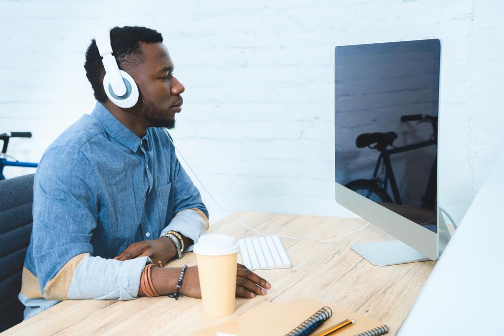 コンピューターを持つテーブルでの作業のヘッドフォンでハンサムなアフリカ系アメリカ人 - 写真・画像