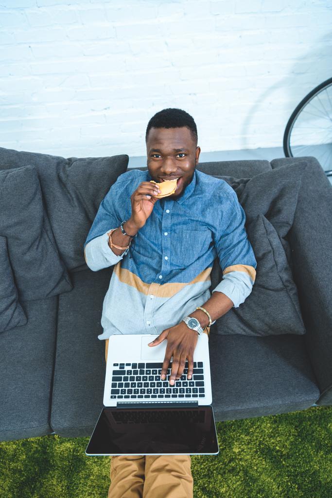 Bello afro-americano che lavora sul computer portatile seduto sul divano e mangia hamburger
 - Foto, immagini