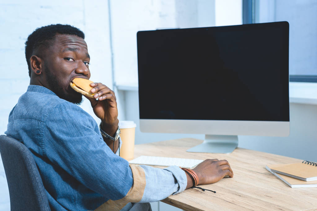 Αφρικανική αμερικανική άνθρωπος που εργάζονται από τον υπολογιστή και να τρώει χάμπουργκερ - Φωτογραφία, εικόνα