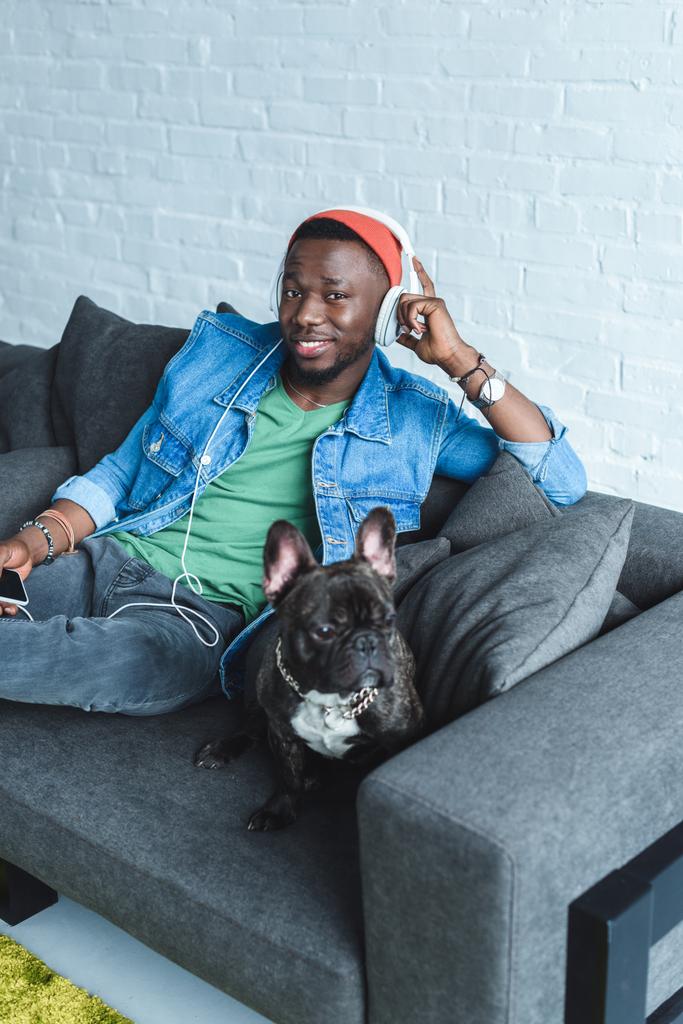 Собака Frenchie на дивані, сидячи афроамериканець людиною в навушниках, прослуховування музики  - Фото, зображення