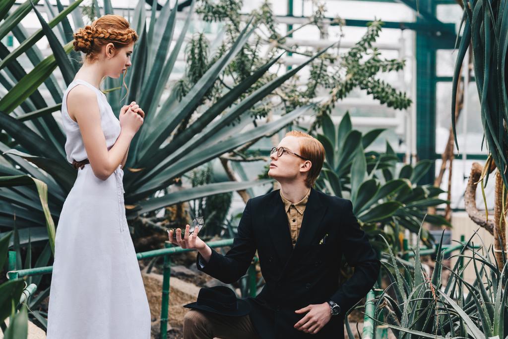 young man standing on knee and making proposal to beautiful redhead girl in botanical garden  - Photo, Image