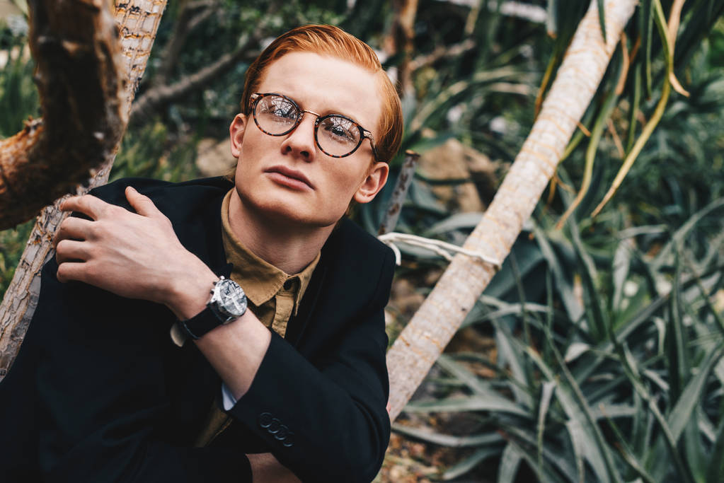 pensive stylish young redhead man in eyeglasses looking away in botanical garden - Photo, Image
