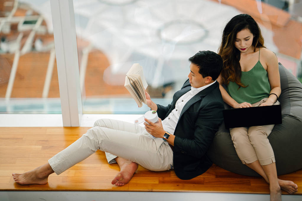 Una pareja asiática china se relaja durante el fin de semana en su apartamento. Una mujer atractiva está trabajando en su portátil en una bolsa de frijoles y su novio está leyendo un libro y apoyándose en ella
. - Foto, imagen