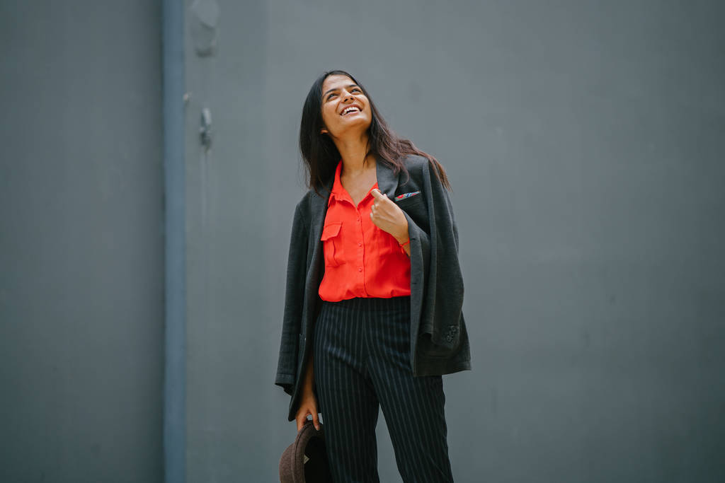 Портрет модной индийской азиатки на сером фоне. Она улыбается и носит умную оранжевую блузку и черные полосатые брюки с пиджаком на плечах
. - Фото, изображение