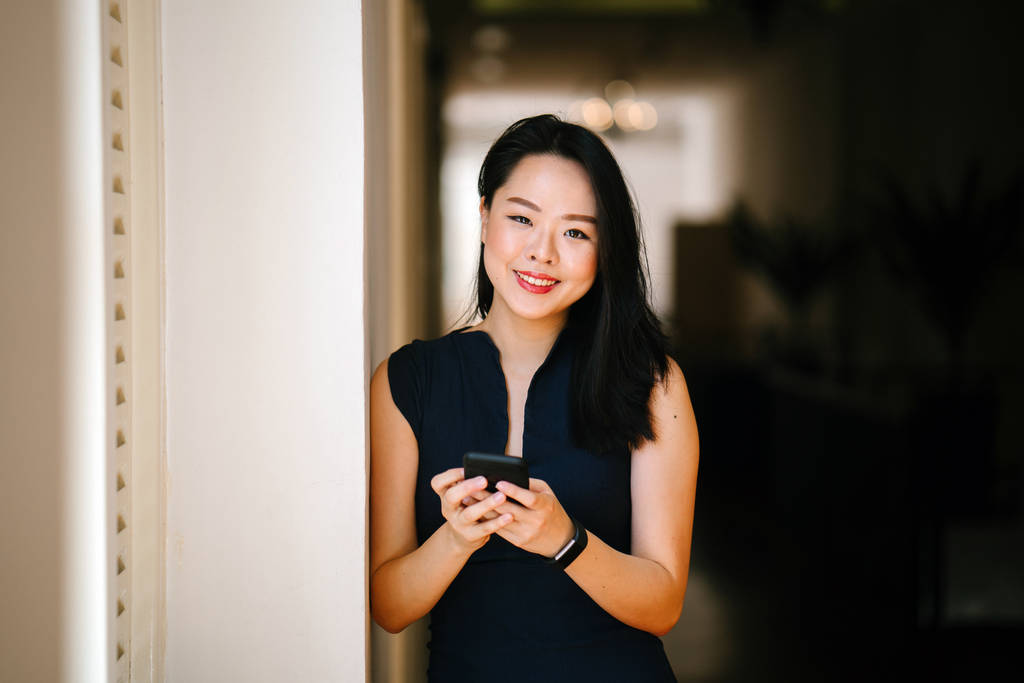 Retrato de una joven y atractiva mujer asiática china usando su teléfono inteligente cerca de una ventana en el día. Ella está usando un vestido azul oscuro profesional y sonriendo
. - Foto, imagen