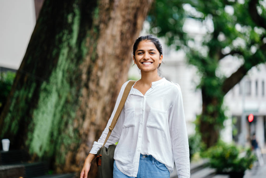 Atrakcyjny i młodych Indian kobiety stojącej obok drzewa w mieście w dzień. Ona jest w casual, białą bluzkę i dżinsy. Istnieje starego budynku w stylu kolonialnym i ruch w tle. - Zdjęcie, obraz