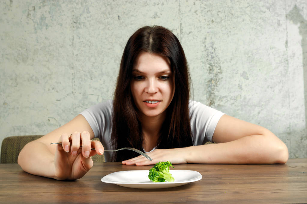 sad young brunette woman dealing with anorexia nervosa or bulimia having small green vegetable on plate. Dieting problems, eating disorder. - Photo, Image