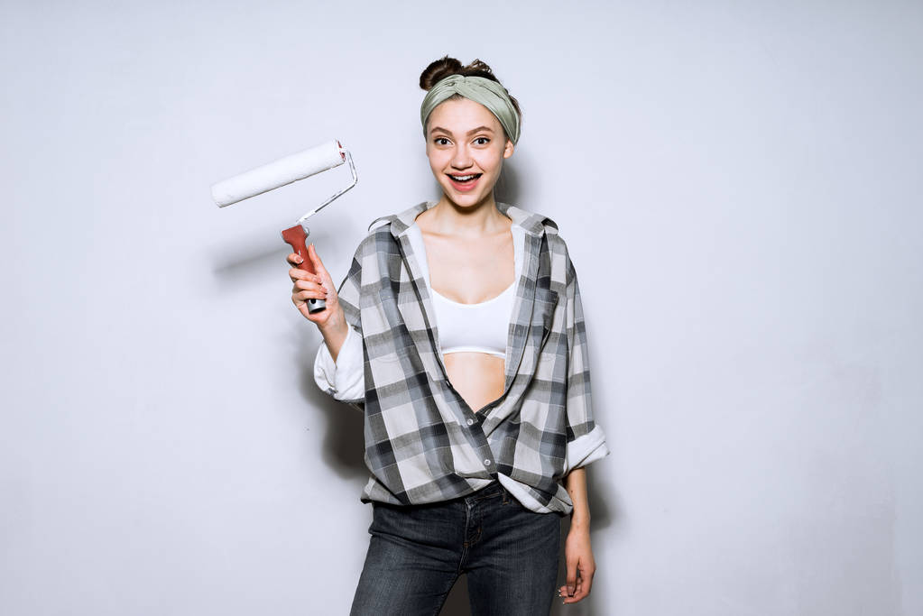 glückliches junges Mädchen im karierten Hemd, das eine Rolle in der Hand hält, um die Wände zu streichen und die Wohnung zu reparieren - Foto, Bild
