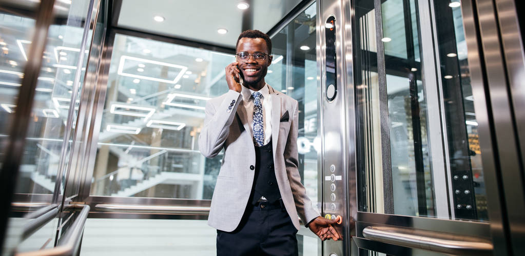 empresario afroamericano en moderno ascensor de cristal hablando por teléfono
 - Foto, imagen
