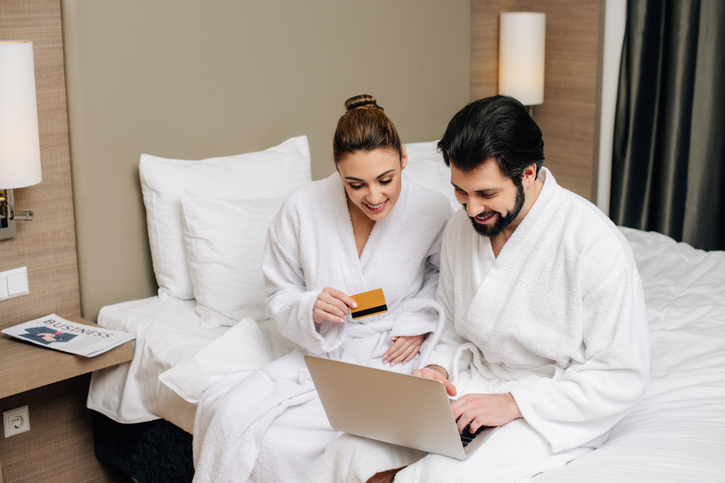 happy couple in bathrobes making e-shopping with laptop and credit card at hotel suite - Photo, Image