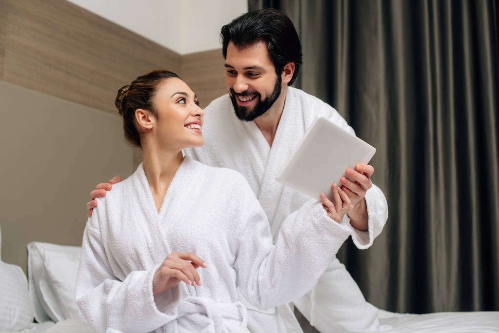 όμορφο ζευγάρι σε μπουρνούζια χρησιμοποιώντας tablet μαζί σε σουίτα ξενοδοχείου - Φωτογραφία, εικόνα