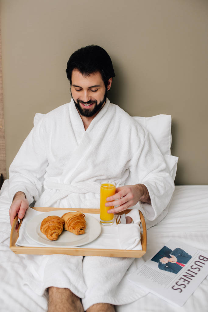 Lächelnder Mann im Bademantel mit Essen auf Tablett auf Bett in Hotelsuite - Foto, Bild