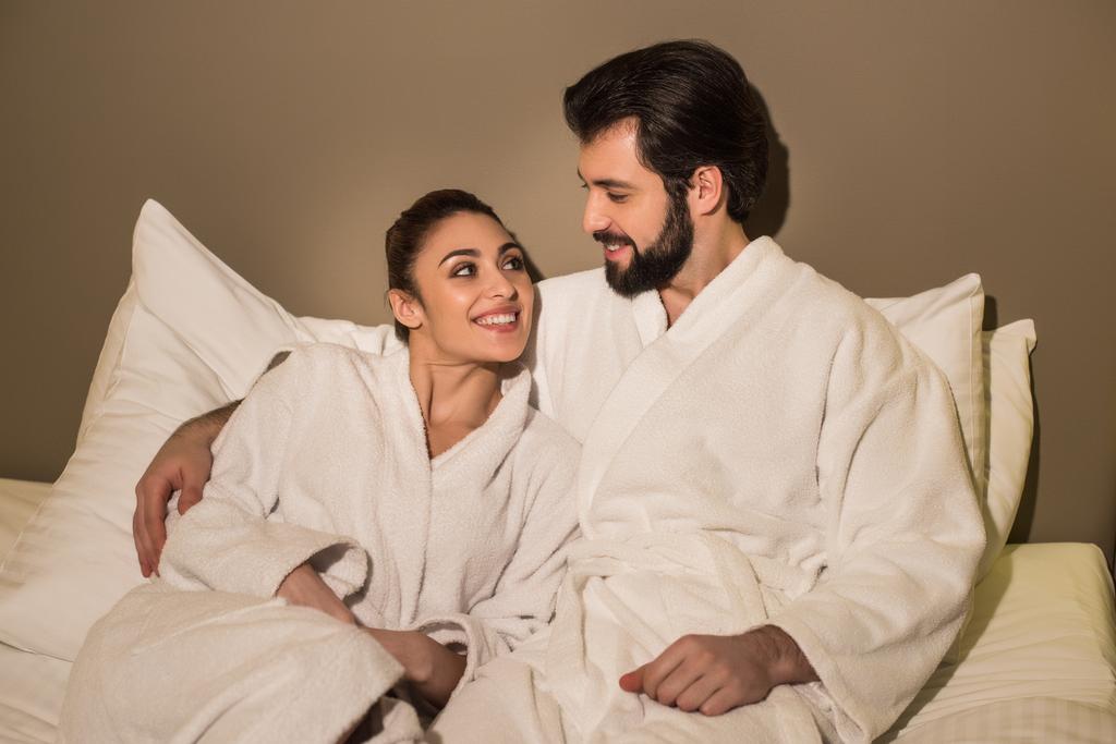hermosa pareja en albornoces abrazándose en la cama de la suite del hotel y mirándose
 - Foto, imagen