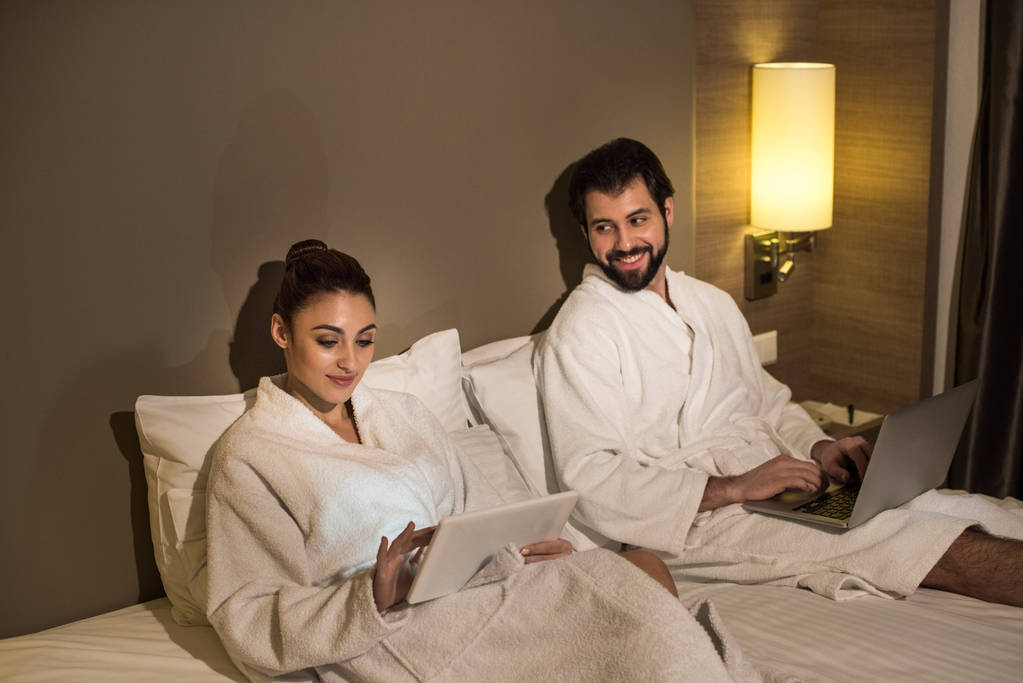 Lächelndes Paar in Bademänteln mit Geräten im Bett der Hotelsuite - Foto, Bild
