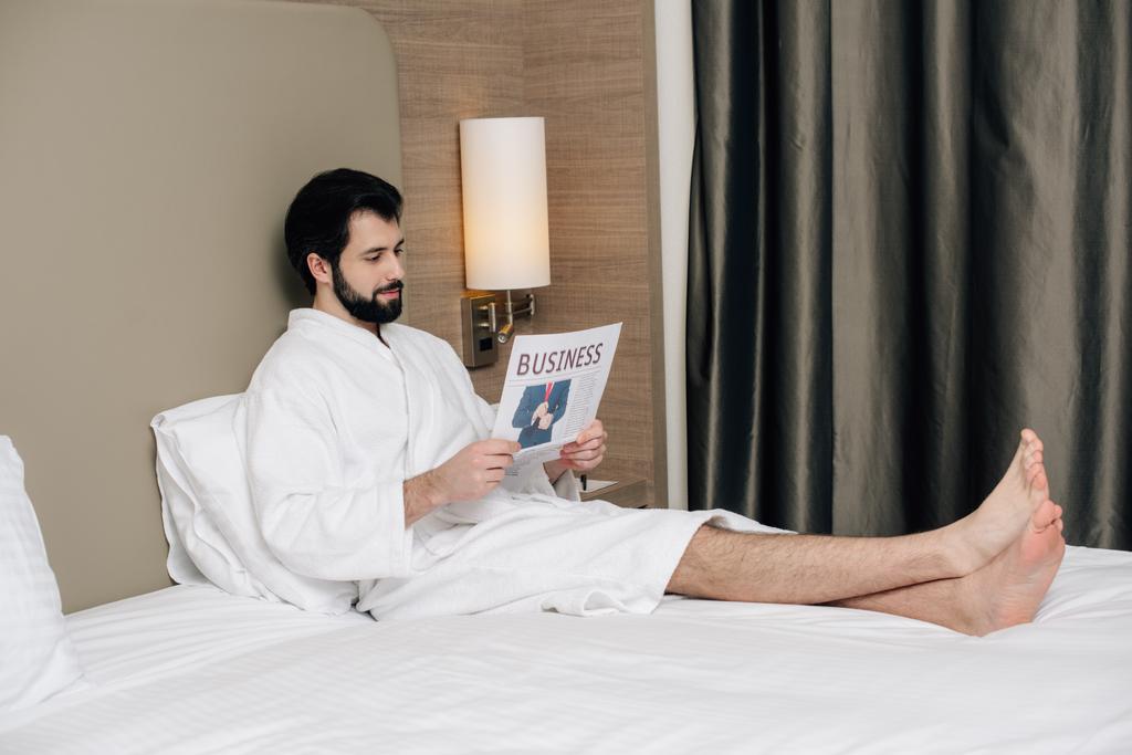 sonriente hombre de negocios en el periódico de lectura de albornoz mientras se relaja en la cama en la suite del hotel
 - Foto, Imagen