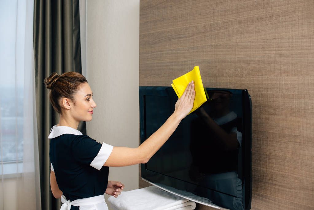 young beautiful maid in uniform cleaning tv screen with rag - Photo, Image