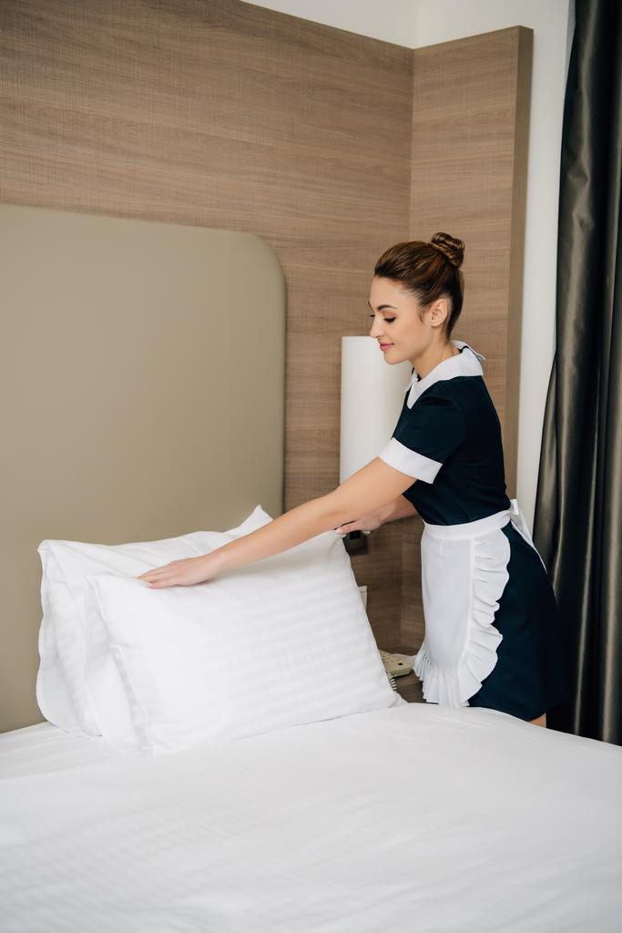 jovem empregada feliz em uniforme fazendo cama na suíte do hotel
 - Foto, Imagem
