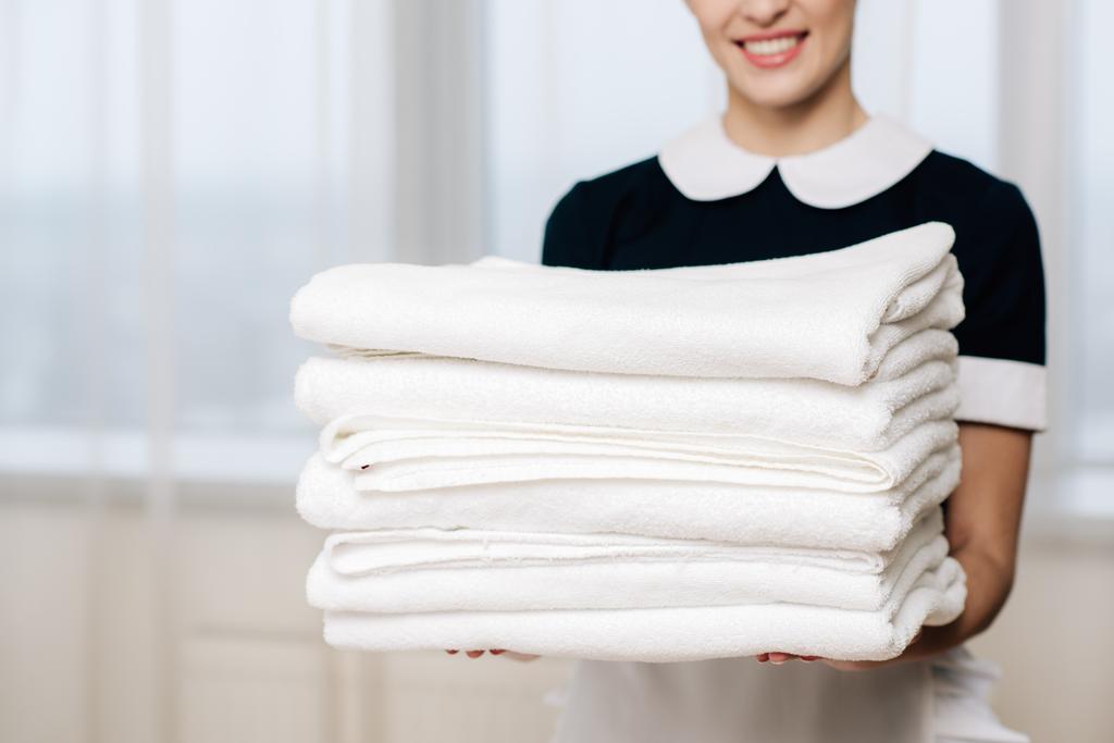 περικοπεί shot από χαμογελαστά υπηρέτρια σε ομοιόμορφη εκμετάλλευση στοίβα από καθαρές πετσέτες - Φωτογραφία, εικόνα