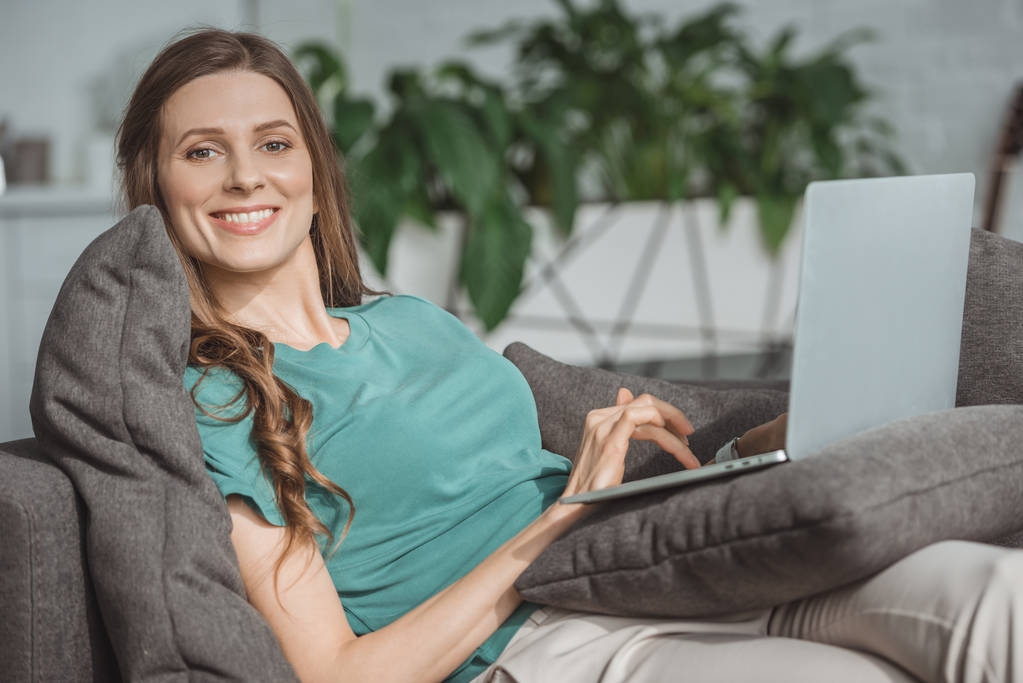 улыбающаяся красивая женщина с помощью ноутбука на диване дома
 - Фото, изображение