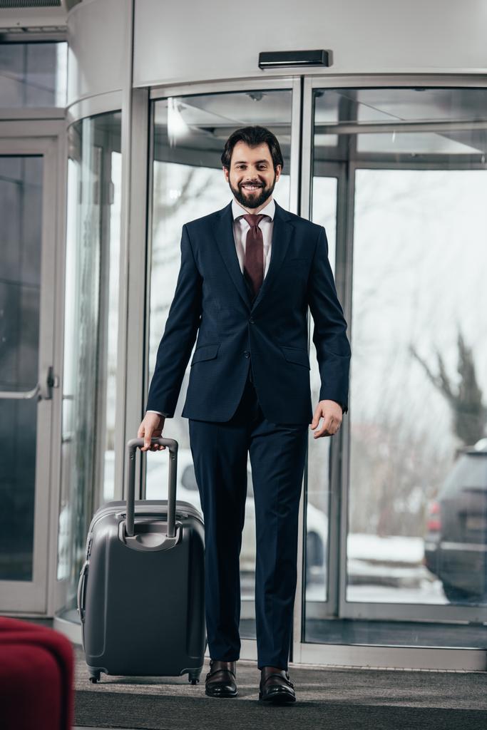 glücklicher gutaussehender Geschäftsmann mit Gepäck geht aus dem Hotel - Foto, Bild