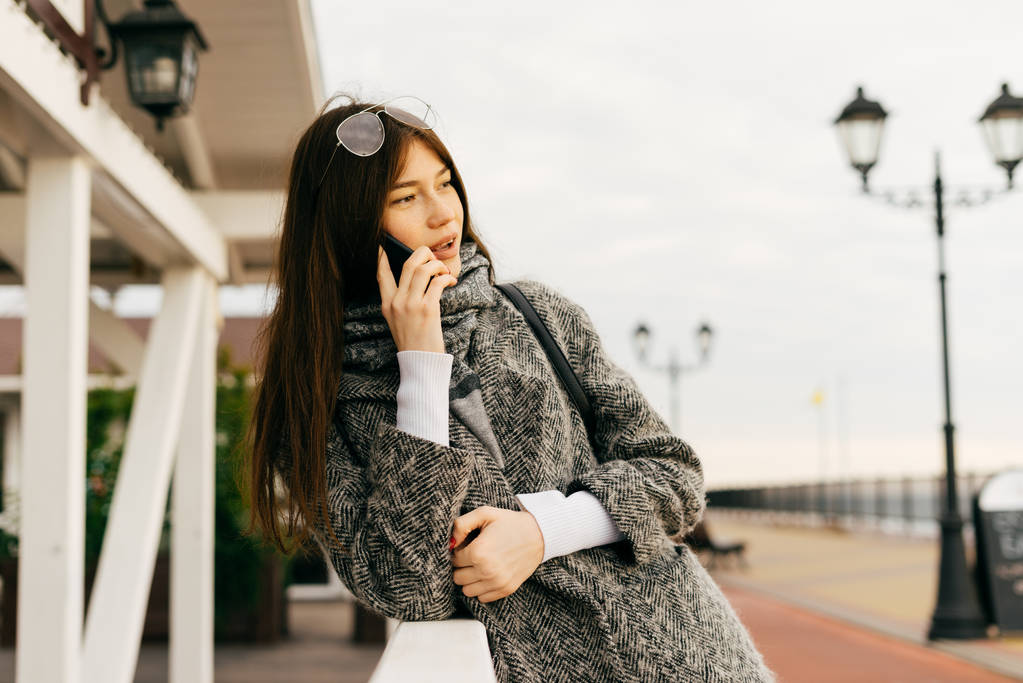 Dziewczyna modnego miasta włosy ciemny szary płaszcz i okulary rozmawia przez telefon, na zewnątrz - Zdjęcie, obraz