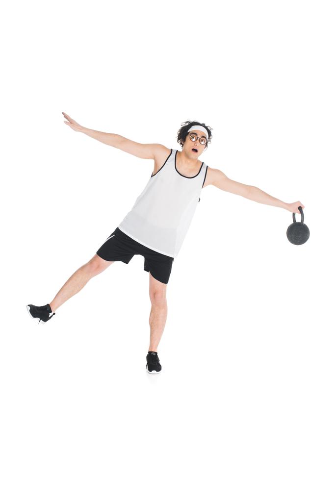 Young skinny sportsman in eyeglasses standing on one leg and holding kettlebell isolated on white - Photo, Image
