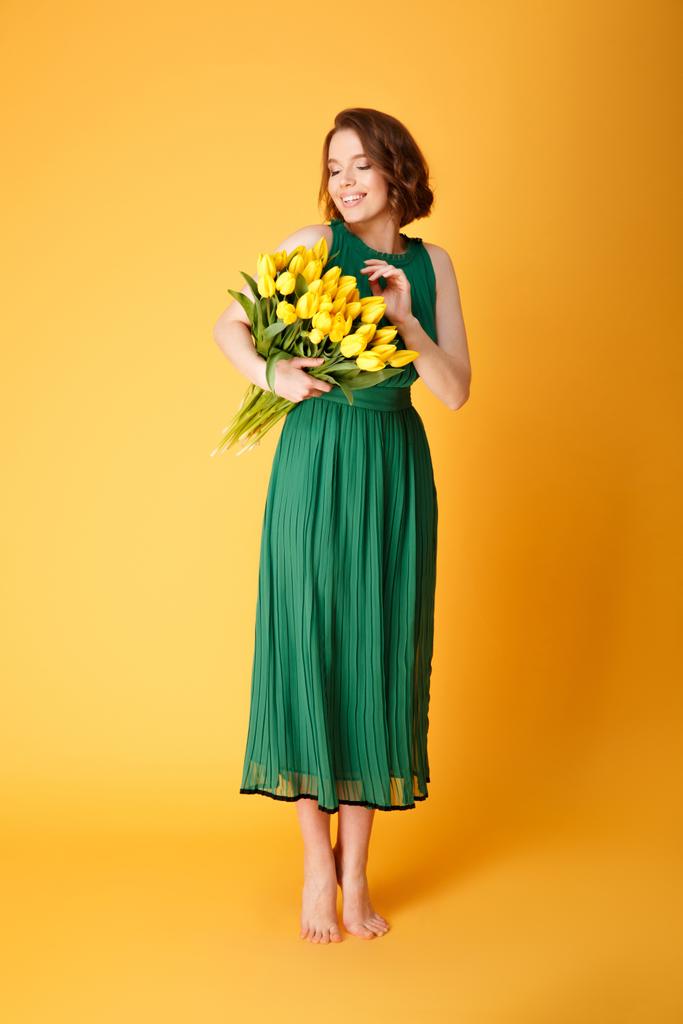 χαμογελαστό γυναίκα στο πράσινο την άνοιξη φόρεμα βλέπουν μπουκέτο των κίτρινων τουλιπών που απομονώνονται σε πορτοκαλί - Φωτογραφία, εικόνα