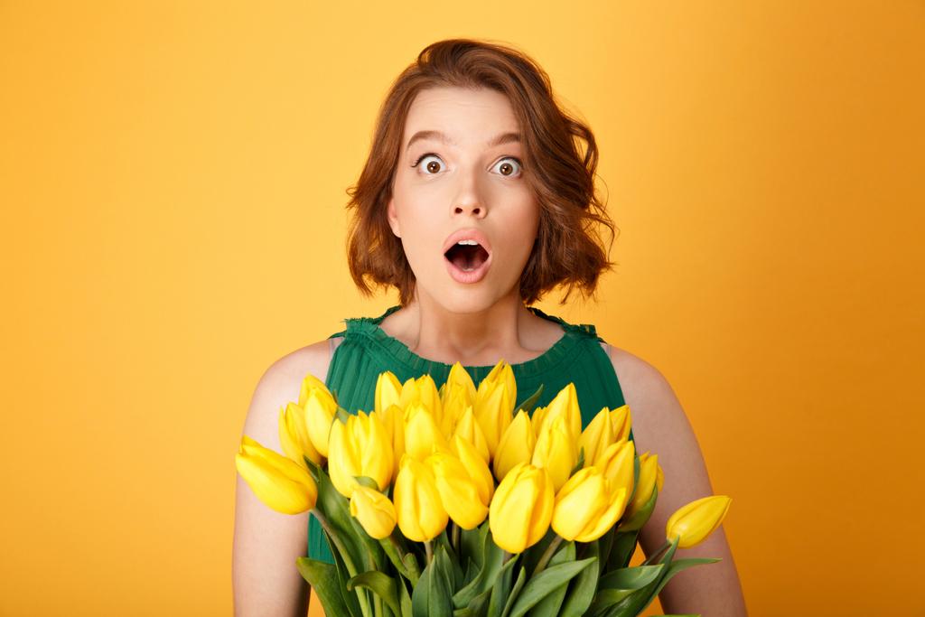 portrait of shocked woman with bouquet of yellow tulips looking at camera isolated on orange - Photo, Image