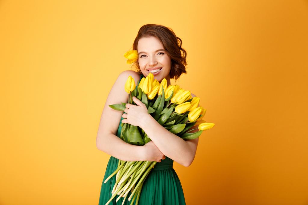 portrait of smiling woman with bouquet of yellow tulips in hands isolated on orange - Photo, Image