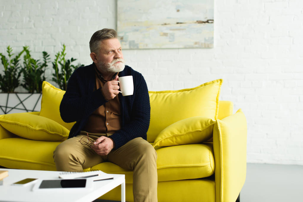 お茶のカップを押しながら黄色のソファに座って、家でよそ見物思いにふける年配の男性  - 写真・画像