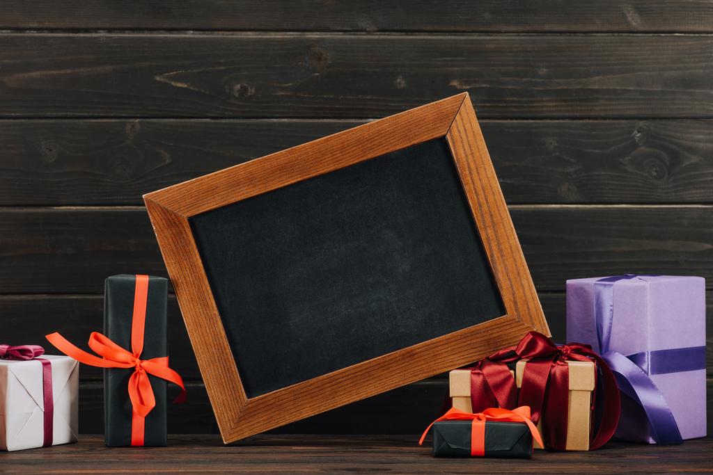 leeg schoolbord in frame met diverse geschenkdozen tegen houten muur - Foto, afbeelding