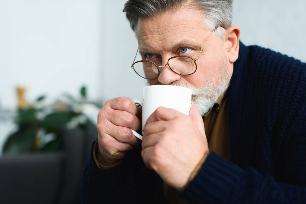 вид крупним планом на старшого чоловіка в окулярах, який п'є з чашки і дивиться вдома
 - Фото, зображення
