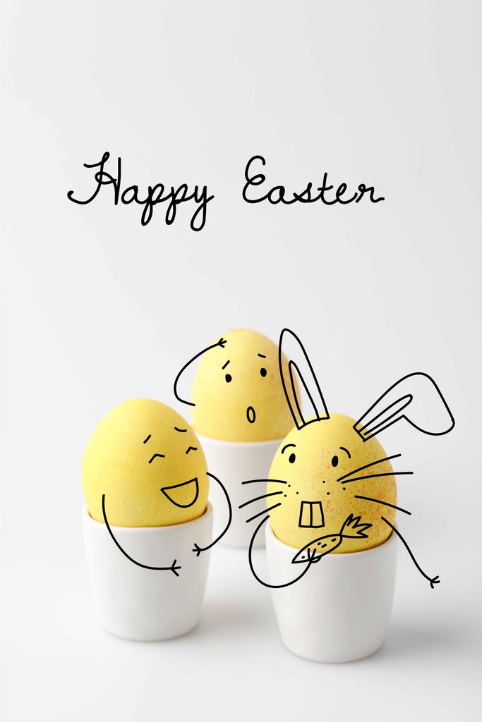 три жовті розмальовані великодні яйця в яєчці стоїть з комічними намальованими обличчями на білому
 - Фото, зображення