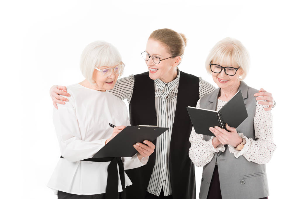 Dos mujeres de negocios con estilo senior que escriben en portapapeles y libro de texto mientras la mujer de negocios madura sostiene sus hombros aislados en blanco
 - Foto, Imagen