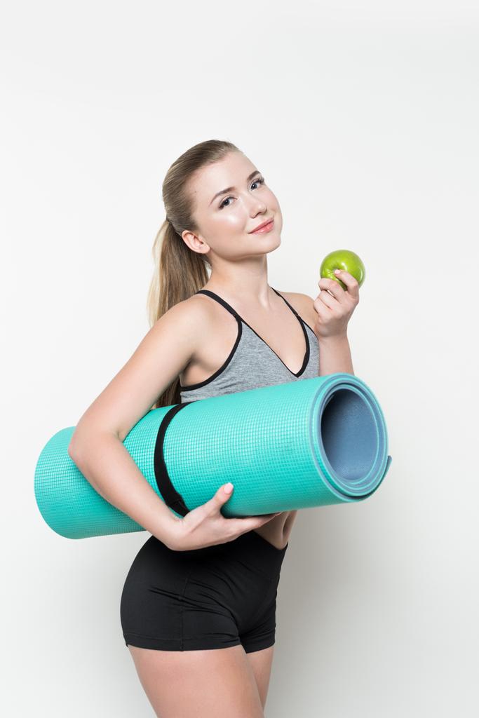 Smiling fitness woman holding apple and yoga mat isolated on white - Photo, Image