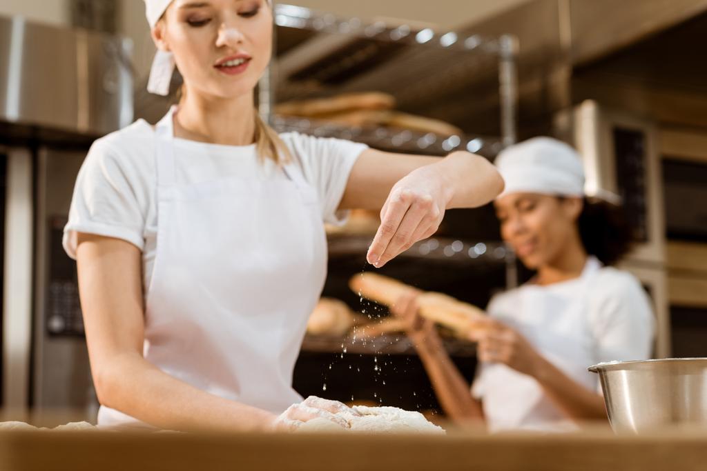Gelukkig vrouwelijke baker kneden deeg en gieten meel daarop bij het bakken van vervaardiging terwijl haar collega die werkt op de achtergrond wazig - Foto, afbeelding