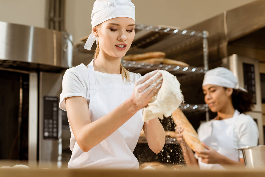 nuori naaras leipuri vaivaamalla taikina leivontaan valmistus, kun hänen kollegansa työskentelee hämärtynyt taustalla
 - Valokuva, kuva