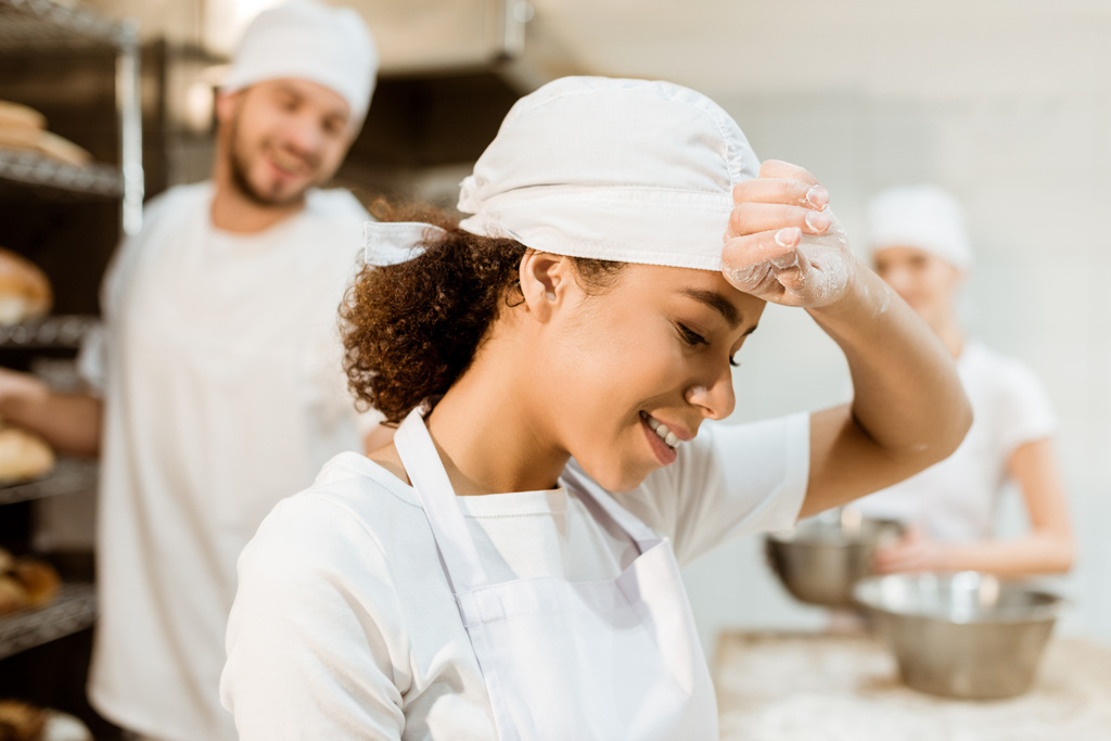 onnellinen ja uupunut nainen leipuri työskentelee leivontaan valmistus, kun hänen kollegansa työskentelevät hämärtynyt taustalla
 - Valokuva, kuva