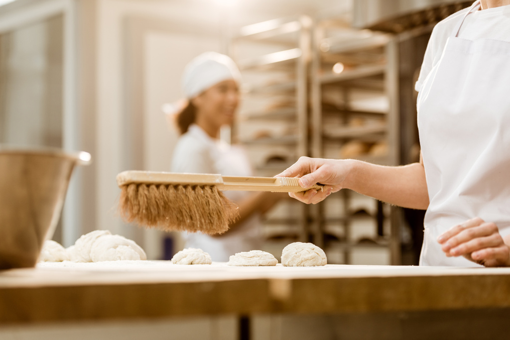 abgeschnittene Aufnahme eines Bäckers, der bei der Backmanufaktur seinen Arbeitsplatz von Mehl mit Pinsel säubert - Foto, Bild