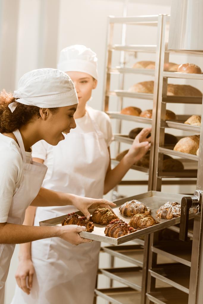 jovens padeiros do sexo feminino colocando pastelaria fresca nas prateleiras na fabricação de panificação
 - Foto, Imagem