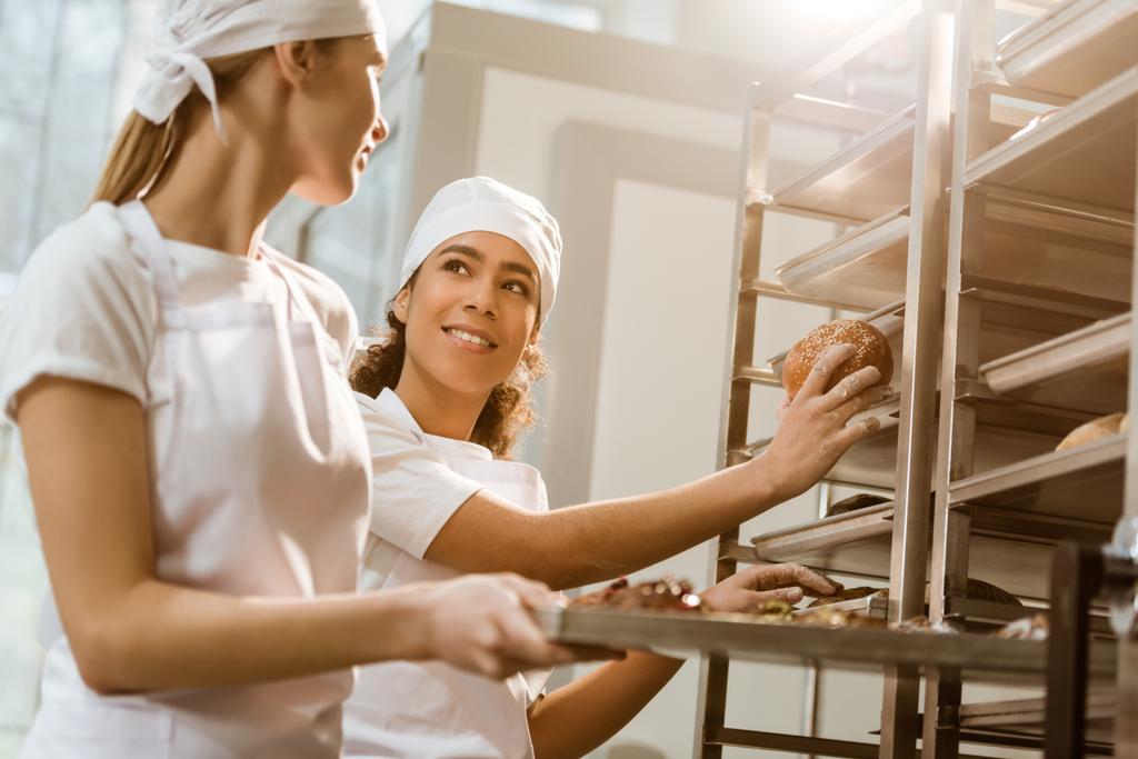 panaderos femeninos felices poniendo pastelería fresca en los estantes en la fabricación de hornear
 - Foto, Imagen