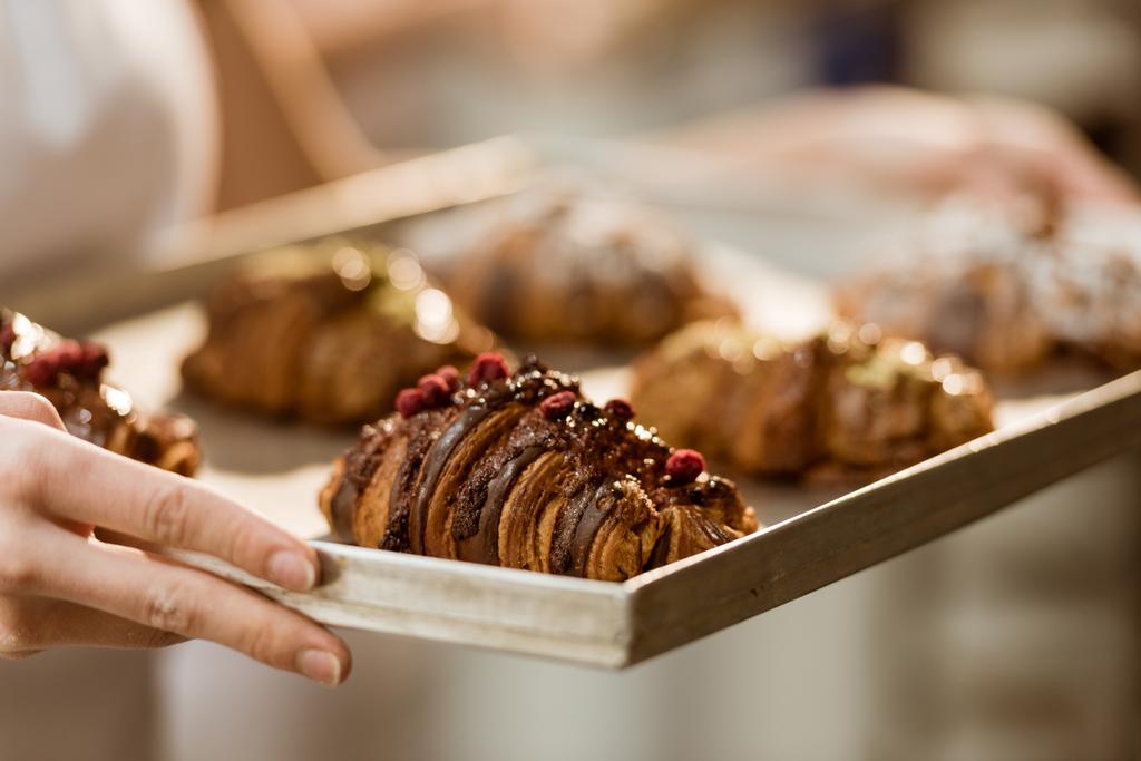 abgeschnittene Aufnahme einer Bäckerin, die Blech mit leckeren Croissants auf Backherstellung hält - Foto, Bild