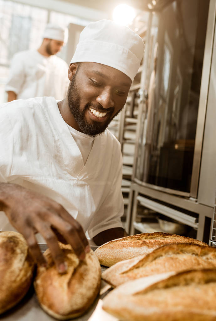 smiling african american baker with tray of fresh loaves of bread on baking manufacture - Photo, Image