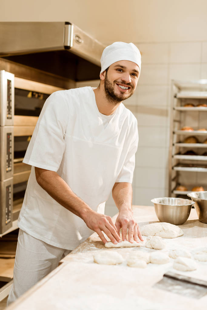 mosolygó fiatal baker nyers tésztát a munkahelyen, a sütés a gyártás előkészítése - Fotó, kép