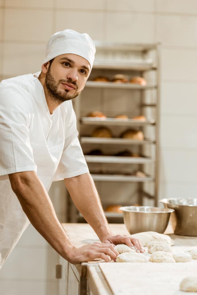 knappe jonge bakker met rauwe deeg op werkplek op bakken vervaardiging - Foto, afbeelding