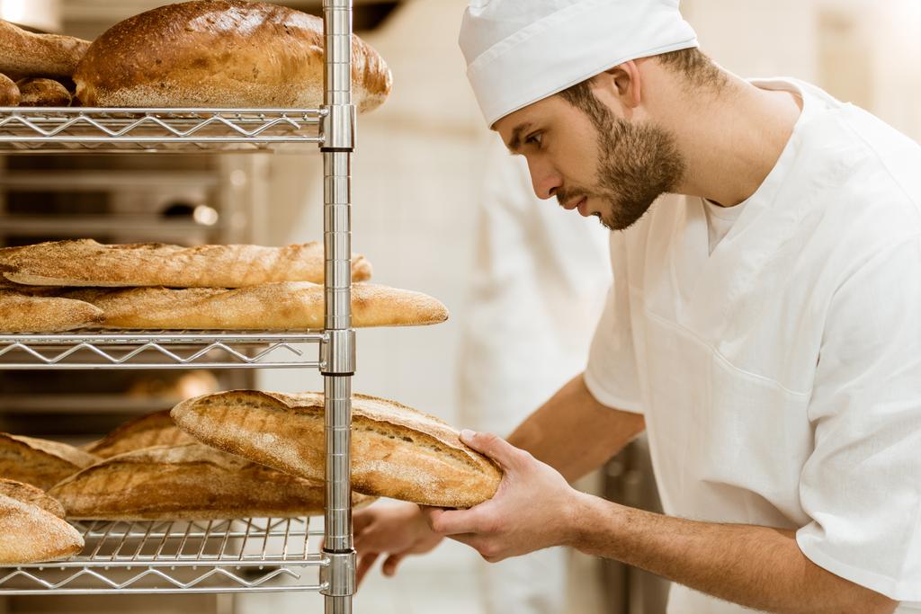 vista lateral do padeiro bonito colocando pão fresco na prateleira na fabricação de cozimento
 - Foto, Imagem