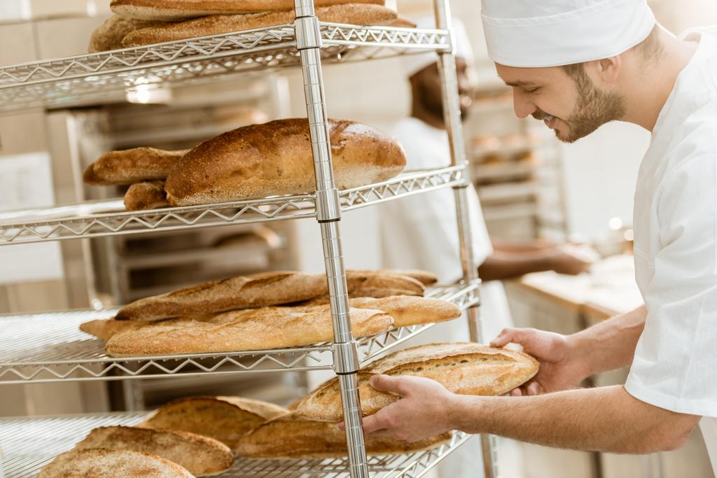 handsome young baker putting fresh bread loaf on shelf at baking manufacture - Photo, Image