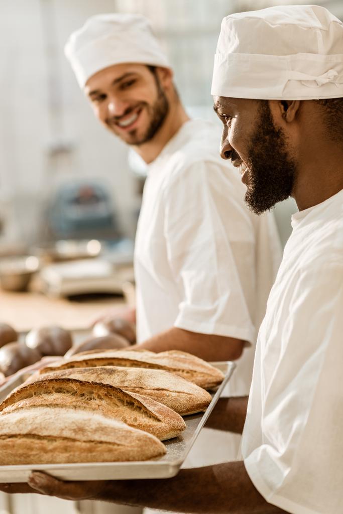 belos jovens padeiros em chapéus na fabricação de panificação olhando para pães frescos na bandeja
 - Foto, Imagem