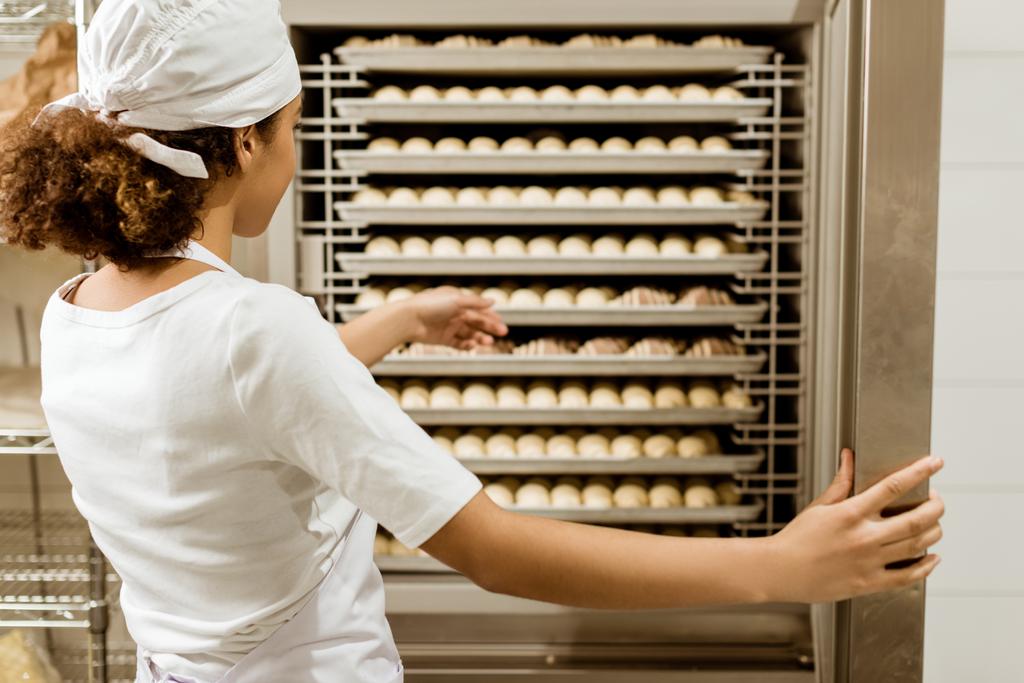 женщина-пекарь указывает на тесто внутри промышленной печи при производстве выпечки
 - Фото, изображение